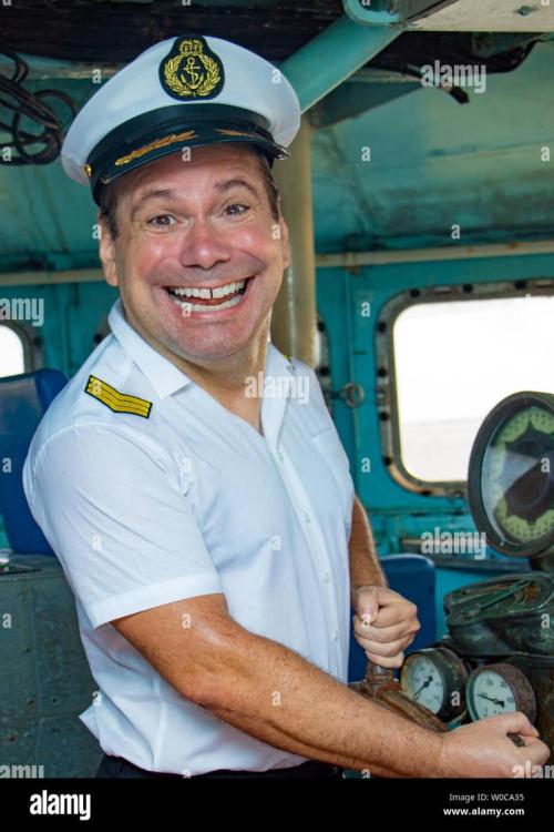 a-laughing-sailor-officer-in-the-uniform-is-steering-the-ship-with-a-rudder-the-funny-captain-standing-in-the-wheelhouse-of-ship-and-looking-to-camer-W0CA35.jpg